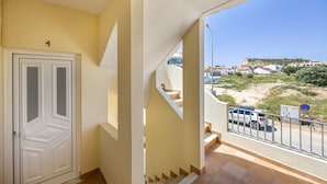 A residential building with a white door, beige walls, and a balcony overlooking a street with parked cars and a hill in the distance. - Apartment Villas Brites nº4 with Shared Pool, Shared Garden, Balcony & Wi-Fi (Burgau)