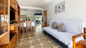 A living room with a sofa, a dining table, and a kitchen area in the background. - Holiday Home Villas Brites nº5 with Open Terrace, Shared Pool & Wi-Fi (Burgau)