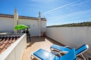 A rooftop terrace with a tiled floor, two blue lounge chairs, a green umbrella, and a building with a clock tower in the background.