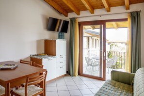 A room with a wooden dining table, chairs, a kitchenette, a flat-screen TV, and a sliding glass door leading to an outdoor area with chairs.