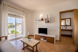 A living room with a TV, a table with a plant, a sliding glass door, and a view of buildings.