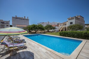 A swimming pool with lounge chairs and an umbrella, surrounded by buildings and greenery.