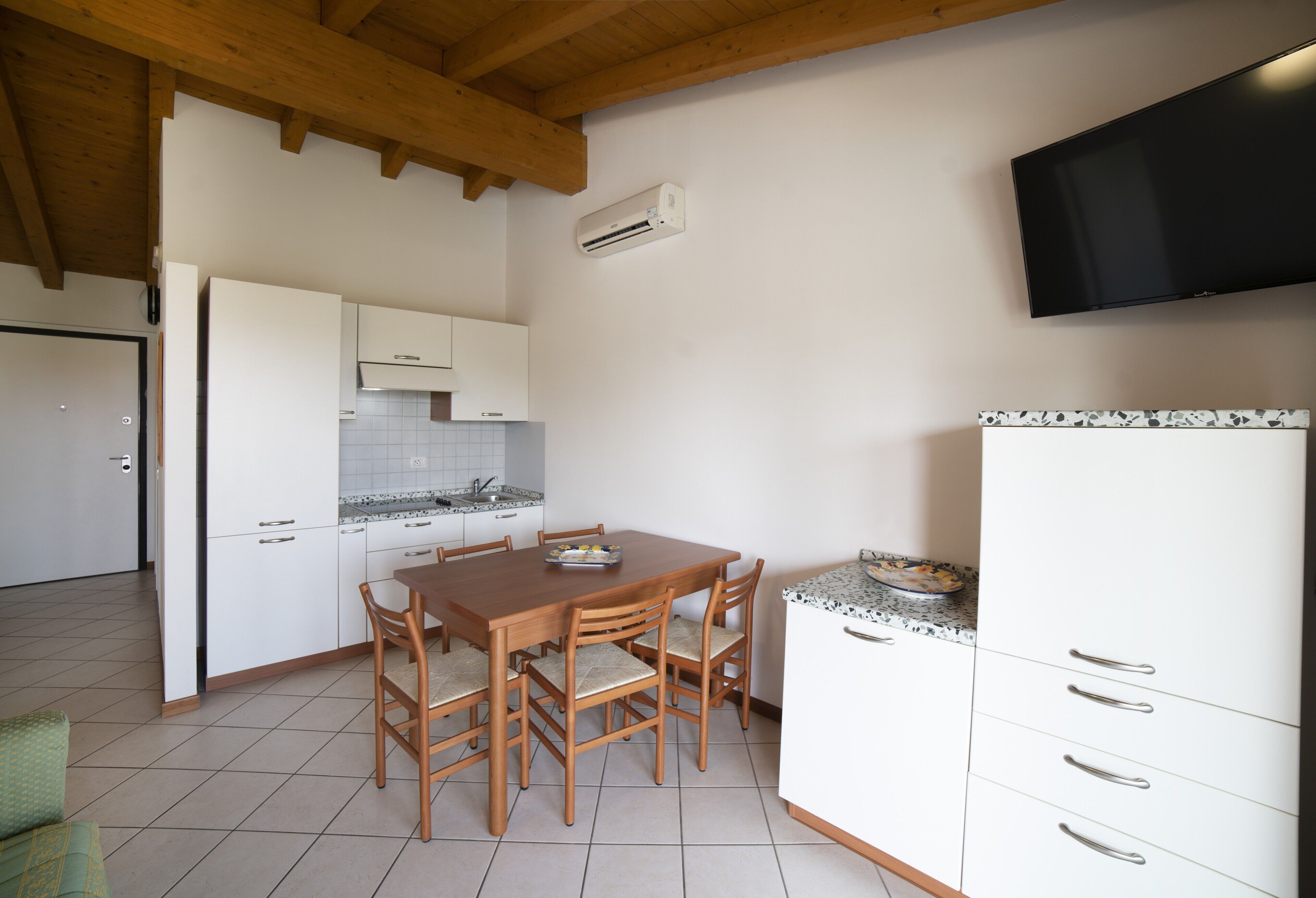 Une cuisine avec une table et des chaises en bois, des armoires blanches, un pare-feu en carreaux et un téléviseur fixé au mur.