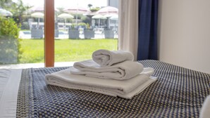 Une pile de serviettes blanches, soigneusement pliées, est posée sur un lit recouvert d’une couvre-lit à motifs. Une fenêtre offre une vue sur des parasols et de la verdure à l’extérieur.