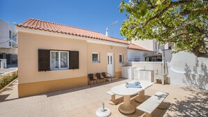 A house with a tiled roof, a patio with a table and chairs, and a small garden area. - Holiday Home Casa Fernandes with Shared Pool, Open Terrace & Wi-Fi (Burgau)
