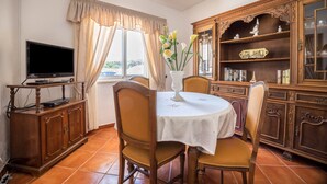 A dining area with a round table, chairs, a television on a cabinet, and a window with curtains. - Holiday Home Casa Fernandes with Shared Pool, Open Terrace & Wi-Fi (Burgau)