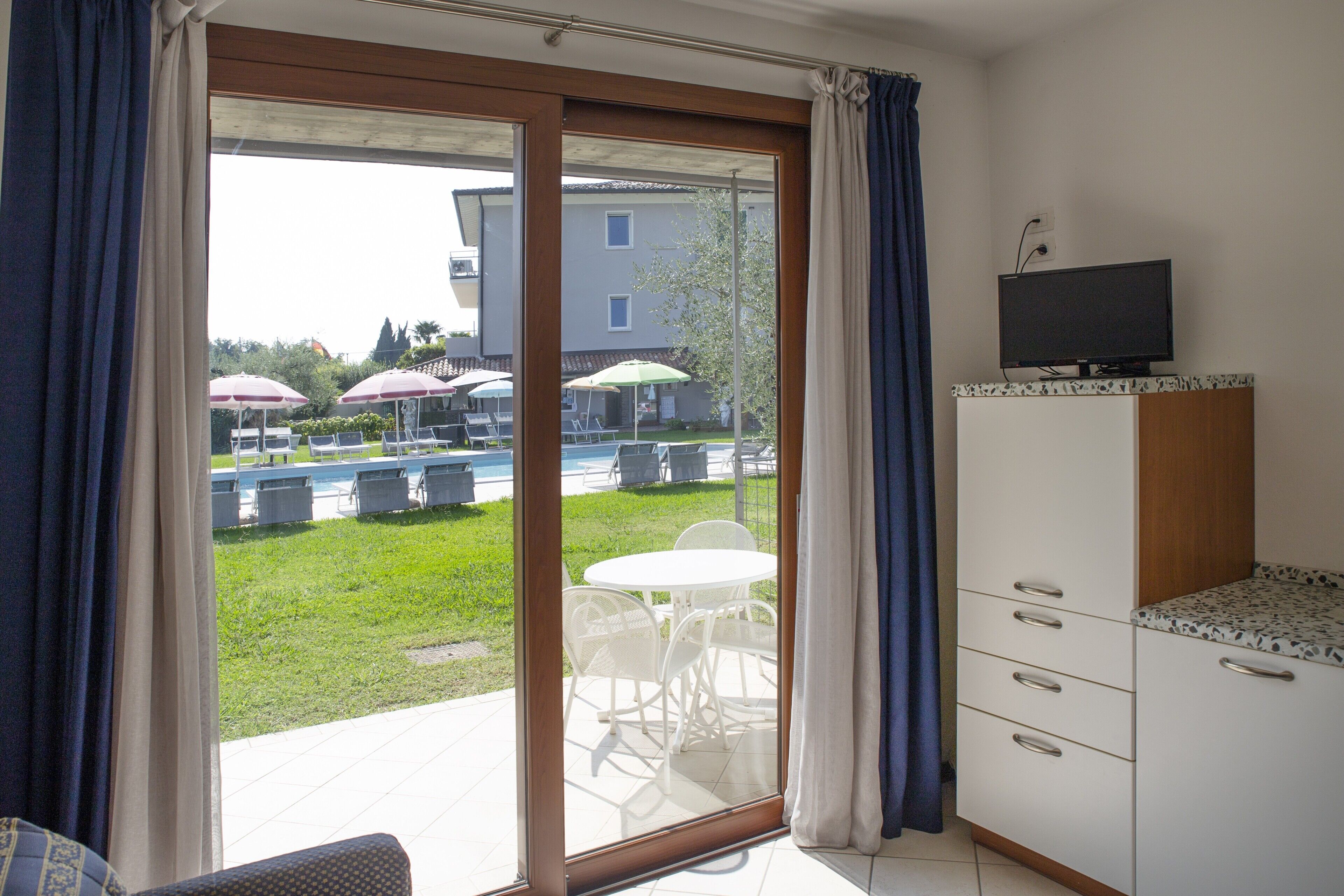 A room with a sliding glass door leading to an outdoor area with a pool and umbrellas, a television on a cabinet, and a small kitchenette.