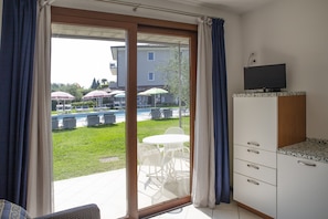 A room with a sliding glass door leading to an outdoor area with a pool and umbrellas, a television on a cabinet, and a small kitchenette.