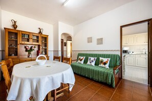 A dining area with a round table, chairs, a green sofa, and a kitchen visible through an open door.