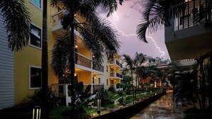 Un edificio de varios pisos con balcones, rodeado de abundante vegetación y un elemento acuático.
