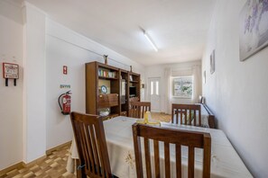 Uma área de refeições com mesa posta para quatro pessoas, cadeiras de madeira, uma estante, um extintor de incêndio e uma janela com cortinas.