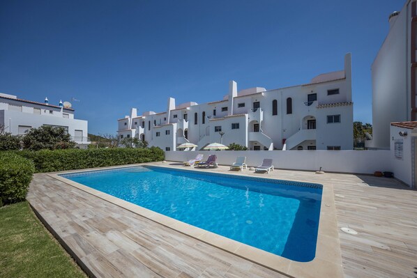 Piscina com espreguiçadeiras e guarda-sóis, cercada por edifícios brancos com varandas.