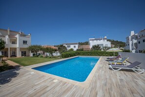 Piscina retangular com um deck ladrilhado, rodeada por edifícios brancos com telhados de terracota, e um céu azul límpido.
