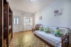 Quarto com guarda-roupa de madeira, cama com colcha branca e almofadas tropicais, cadeira de madeira e janela com cortinas.