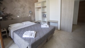 Cama bem arrumada com colcha cinza e lençóis brancos, mesa de cabeceira com abajur, guarda-roupa e parede com padrão floral.