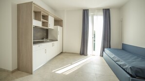 A compact room with a kitchenette, a sofa, and a window with curtains. - Deluxe Suite 1 with Shared Pool and Private Balcony (Manerba del Garda)