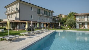 Une piscine avec des chaises longues et des parasols, un bâtiment doté de balcons et un espace jardin.