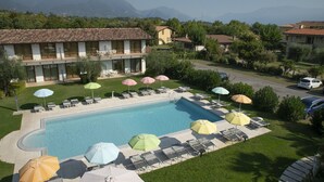 A swimming pool with lounge chairs and umbrellas, surrounded by greenery and a view of mountains. - Deluxe Suite 1 with Shared Pool and Private Balcony (Manerba del Garda)