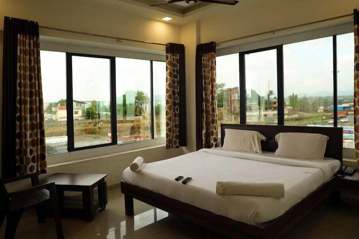 A hotel room with a large bed, a small table, and a view of the city through large windows.