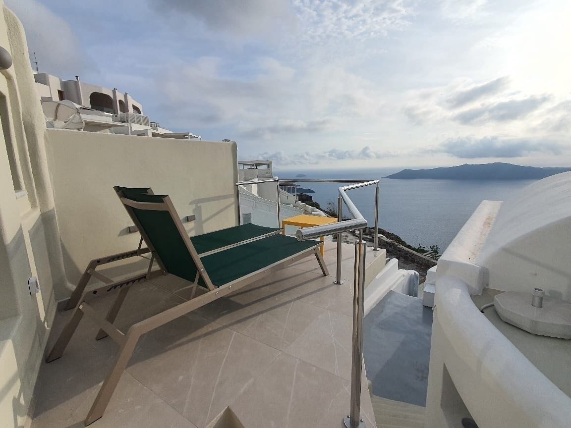 Un balcon avec une chaise verte, une petite table et une vue sur la mer et les montagnes lointaines.