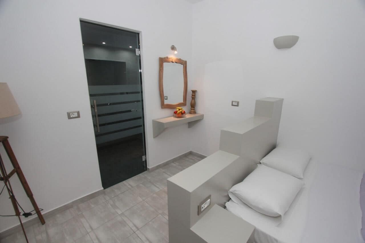 A modern bathroom with a glass door, a wooden-framed mirror, a small shelf with decorative items, and a white bench with pillows.