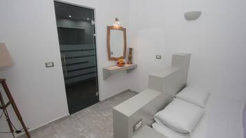 A modern bathroom with a glass door, a wooden-framed mirror, a small shelf with decorative items, and a white bench with pillows.