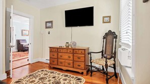 Quarto com cômoda de madeira, uma cadeira, televisão na parede e tapete estampado no chão.