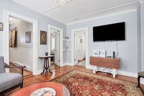 Een woonkamer met een flatscreen televisie aan de muur, een houten salontafel, een kamerbreed tapijt met patroon en een grijze fauteuil.