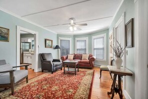 A living room with a sofa, armchair, coffee table, and a dining table with chairs. There is a ceiling fan, a window with blinds, and a door leading to another room.