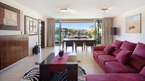 Een moderne woonkamer met een rode bank, een glazen salontafel en een zebrapattend tapijt. Er is een zithoek met een tafel en stoelen, en een balkon met uitzicht op palmbomen.