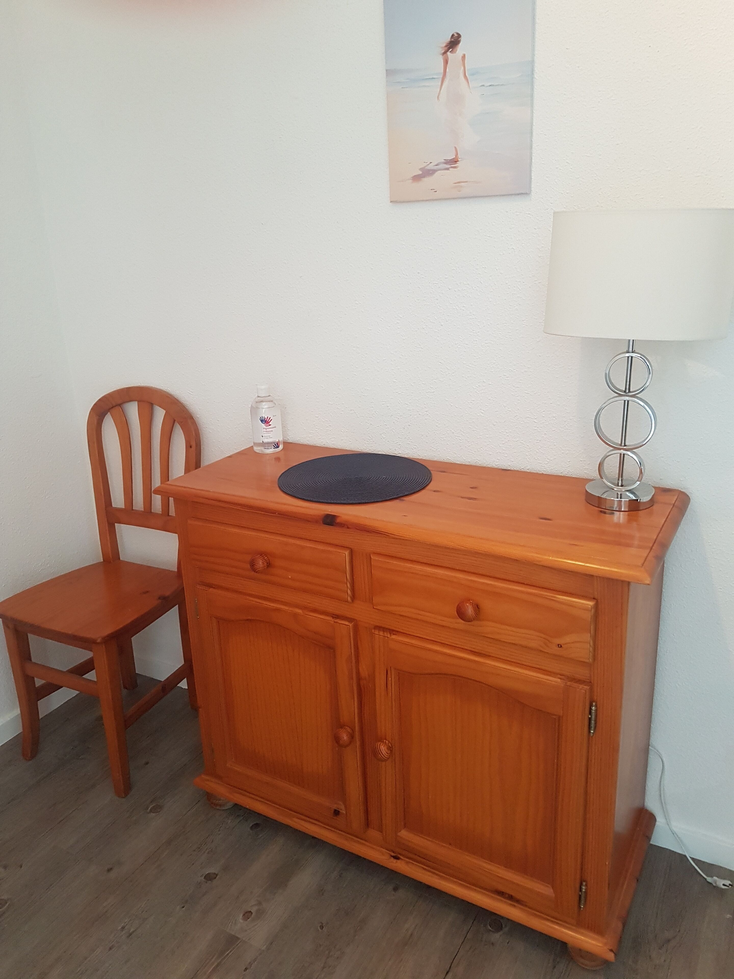 Een houten kast met twee lades en een ronde mat erop, een houten stoel, een fles handdesinfecterend middel, een lamp en een schilderij aan de muur.