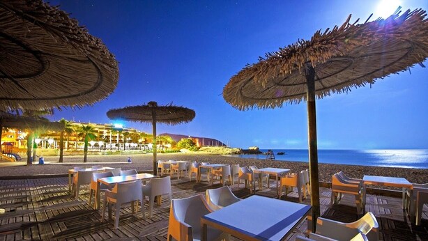 Een buitenterras met rieten parasols, houten tafels en stoelen, met uitzicht op een strand en zee in de schemering.