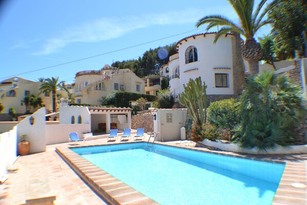 A swimming pool with lounge chairs, surrounded by buildings with Mediterranean architecture, palm trees, and a clear sky.