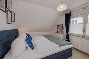 Un dormitorio con una cama, una mesita de noche, una ventana con cortinas y cuadros colgados en la pared.