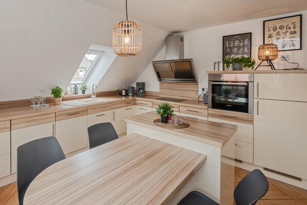 A modern kitchen with a wooden countertop, a dining table, and chairs. There is a built-in oven and a coffee machine.