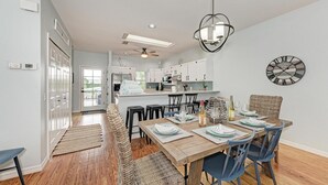 A modern kitchen with white cabinets, a central island, and a dining area with a wooden table and blue chairs. - Aloha Fern: North End Beach House, Pool by Anna Maria Life Vacation Rentals (Anna Maria)