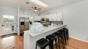 A modern kitchen with white cabinets, a stainless steel refrigerator, a ceiling fan, and a bar area with stools. - Aloha Fern: North End Beach House, Pool by Anna Maria Life Vacation Rentals (Anna Maria)