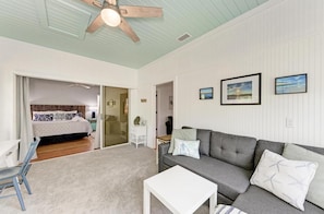 A spacious living area with a grey sofa, a white coffee table, and a ceiling fan. A bedroom with a bed and a bathroom with a shower and toilet are visible through an open door.
