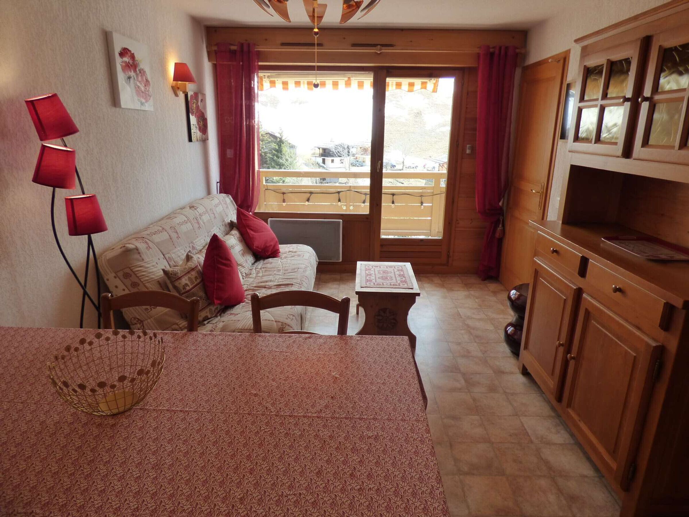 A cozy room with a sofa, a small table, and a TV. There's a window with curtains, a wooden bench outside, and a wooden cabinet.
