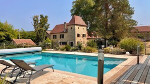 Une piscine entourée de chaises longues, un bâtiment en pierre avec un toit rouge et une végétation luxuriante.