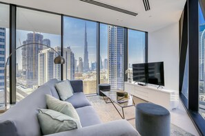 Un salon moderne avec un grand canapé, une table basse et un téléviseur. La pièce est dotée de grandes fenêtres du sol au plafond qui offrent une vue sur la ville.