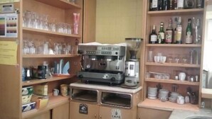 A compact kitchen with wooden cabinets, a coffee machine, and shelves stocked with glassware and bottles.