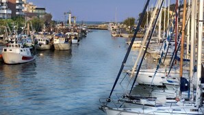 A marina with boats docked along the water, buildings on the left, and a clear sky.