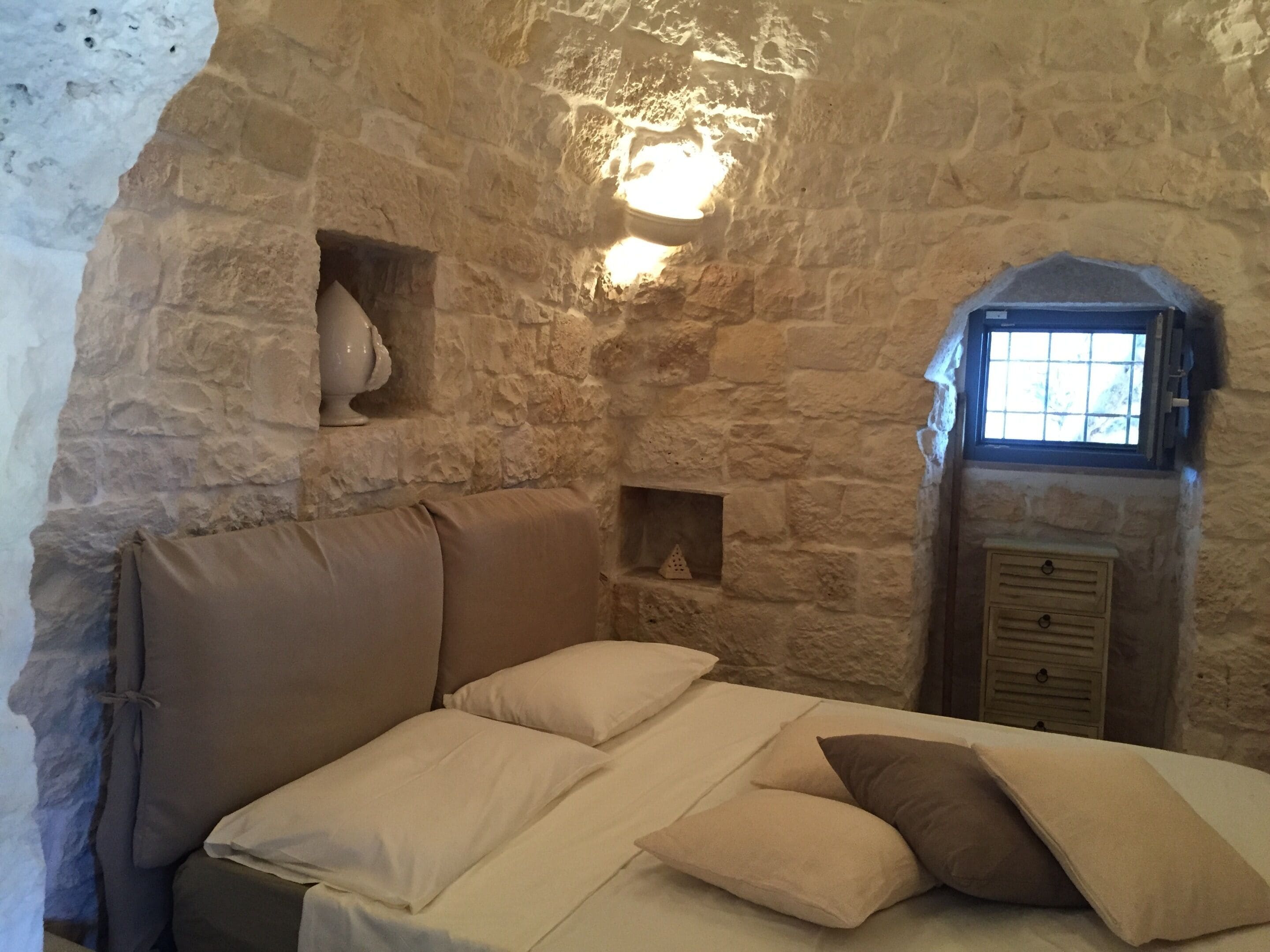 A stone-walled room with a bed, a small window, and a wall-mounted light fixture.