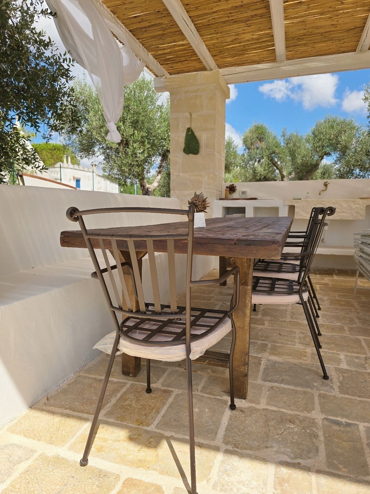 An outdoor patio with a wooden table and metal chairs, stone flooring, and a pergola overhead.
