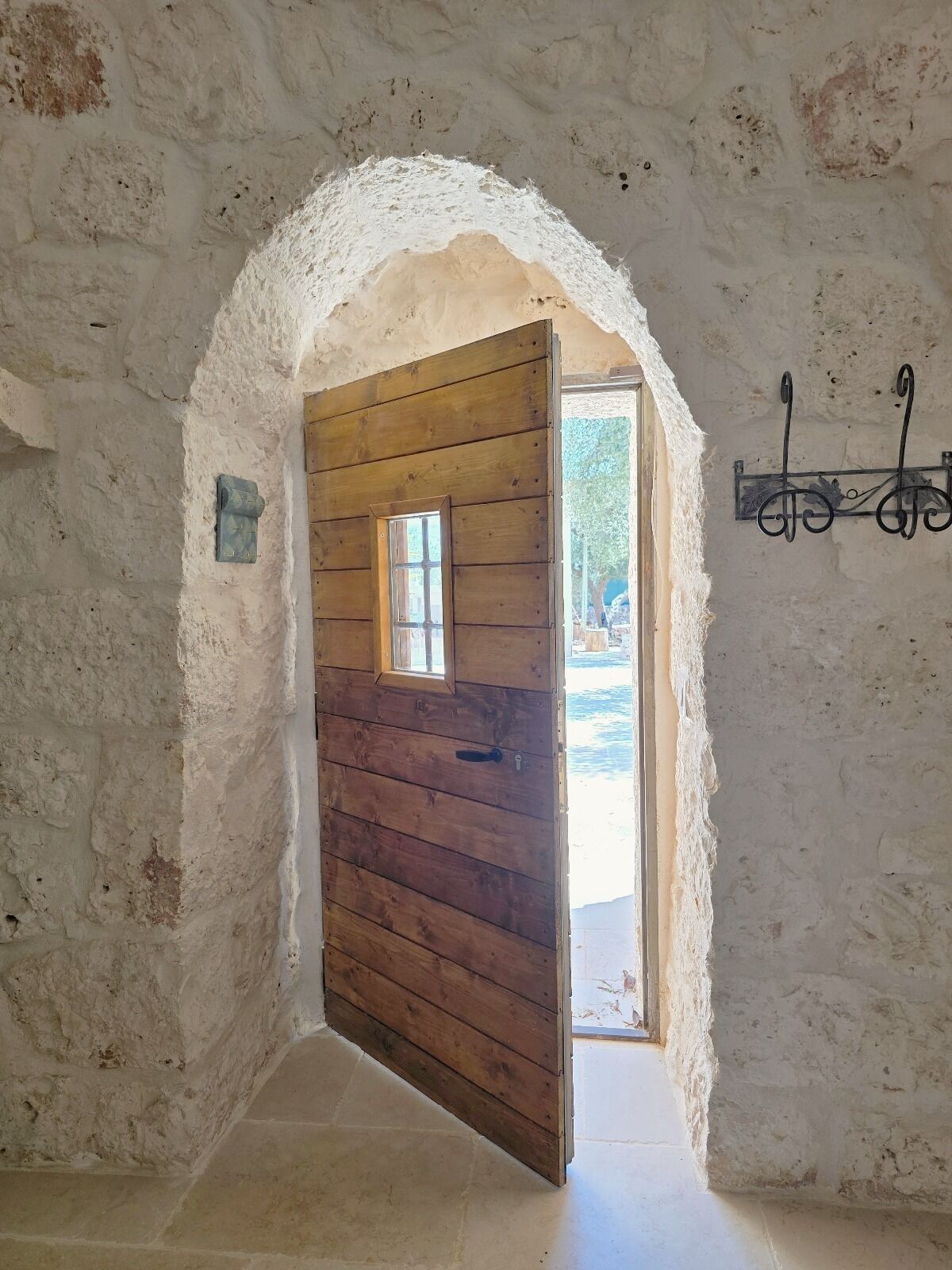 Eine rustikale Holztür mit kleinem Fenster, die in einen Raum mit Steinwand und Metallleuchte führt.
