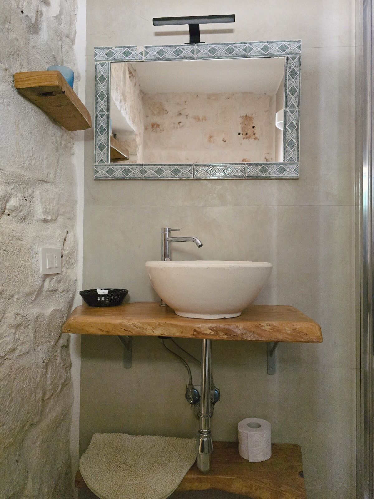 A bathroom with a white sink, a mirror with a decorative frame, a wooden shelf, and a towel on the floor.