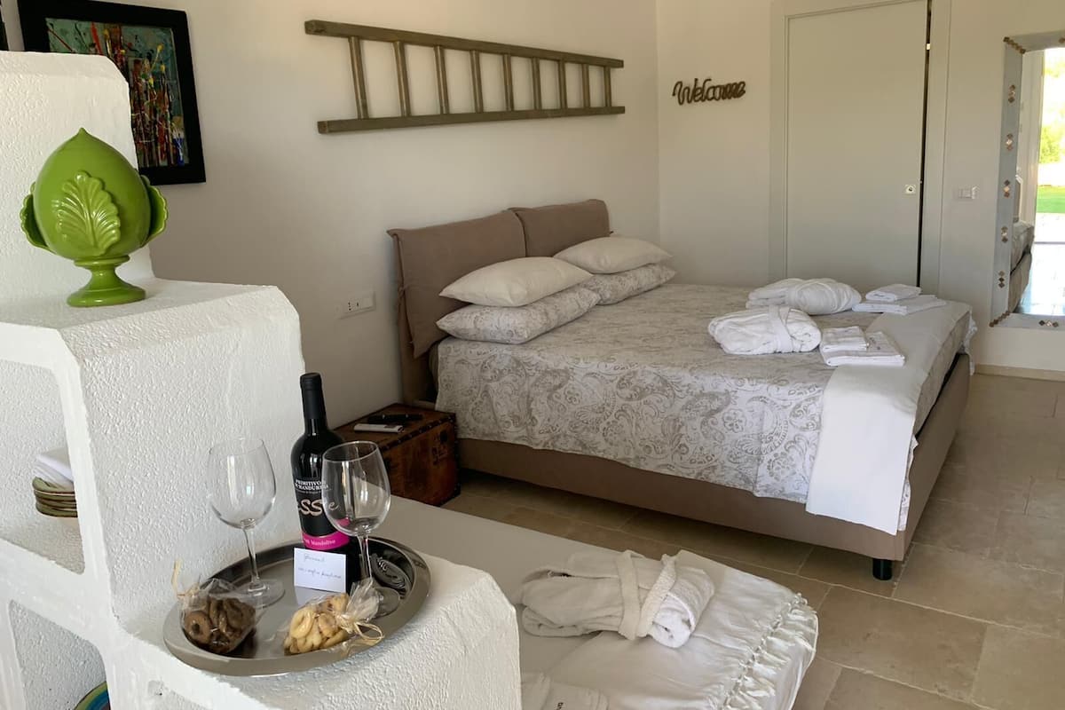 A hotel room with a bed, a bedside table with a wine bottle and glass, a tray with snacks, and a green decorative item.