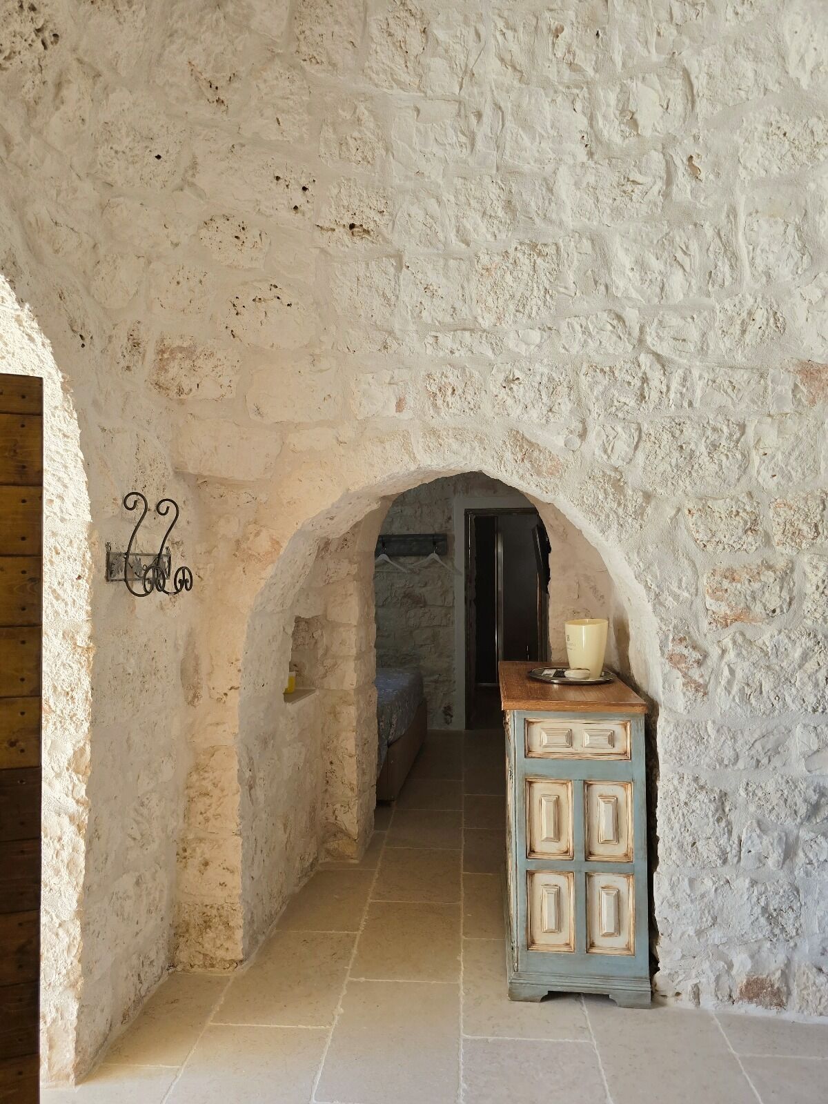 Un estrecho pasillo de piedra con una rústica puerta de madera a la izquierda y una pequeña mesa con una vela a la derecha.