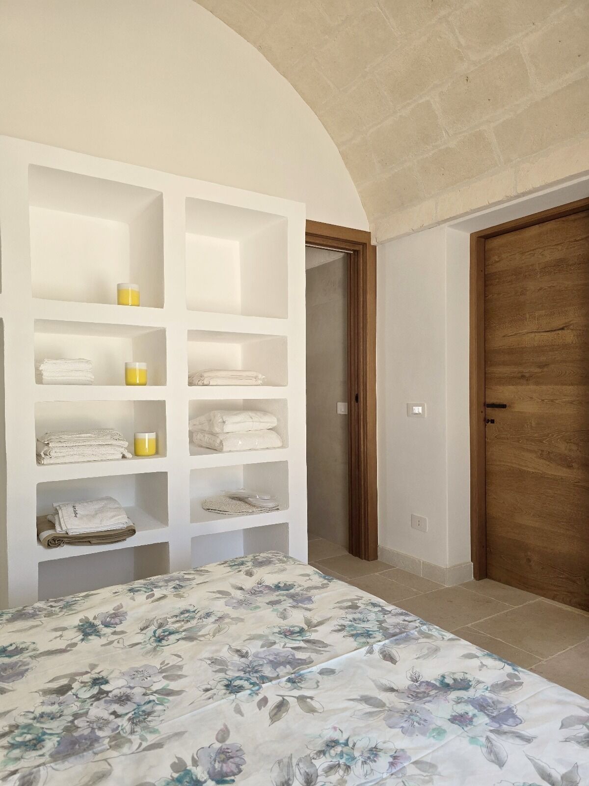 Une chambre avec un lit, une porte en bois et une étagère intégrée où sont rangées des serviettes pliées et des bougies.